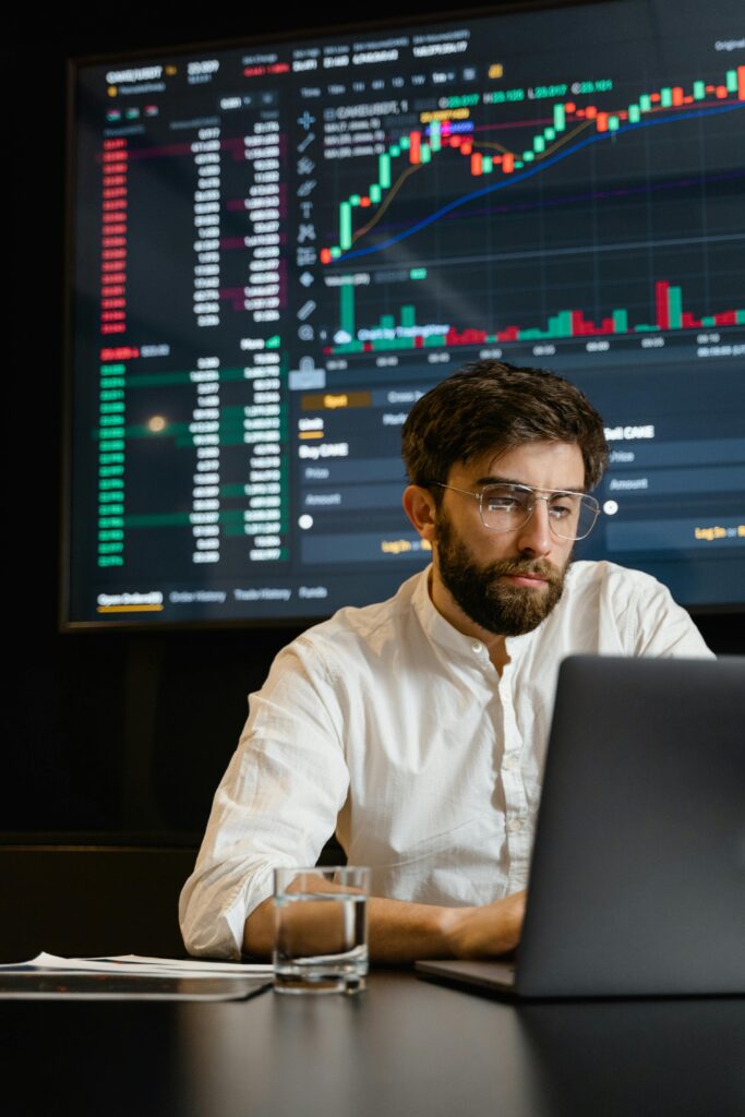 A man analyzing cryptocurrency trends on a laptop with trading graphs displayed on a screen.
